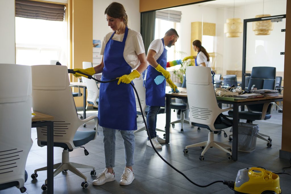 Trzej pracownicy sprzątający biuro, jeden z nich odkurza krzesło parownicą.